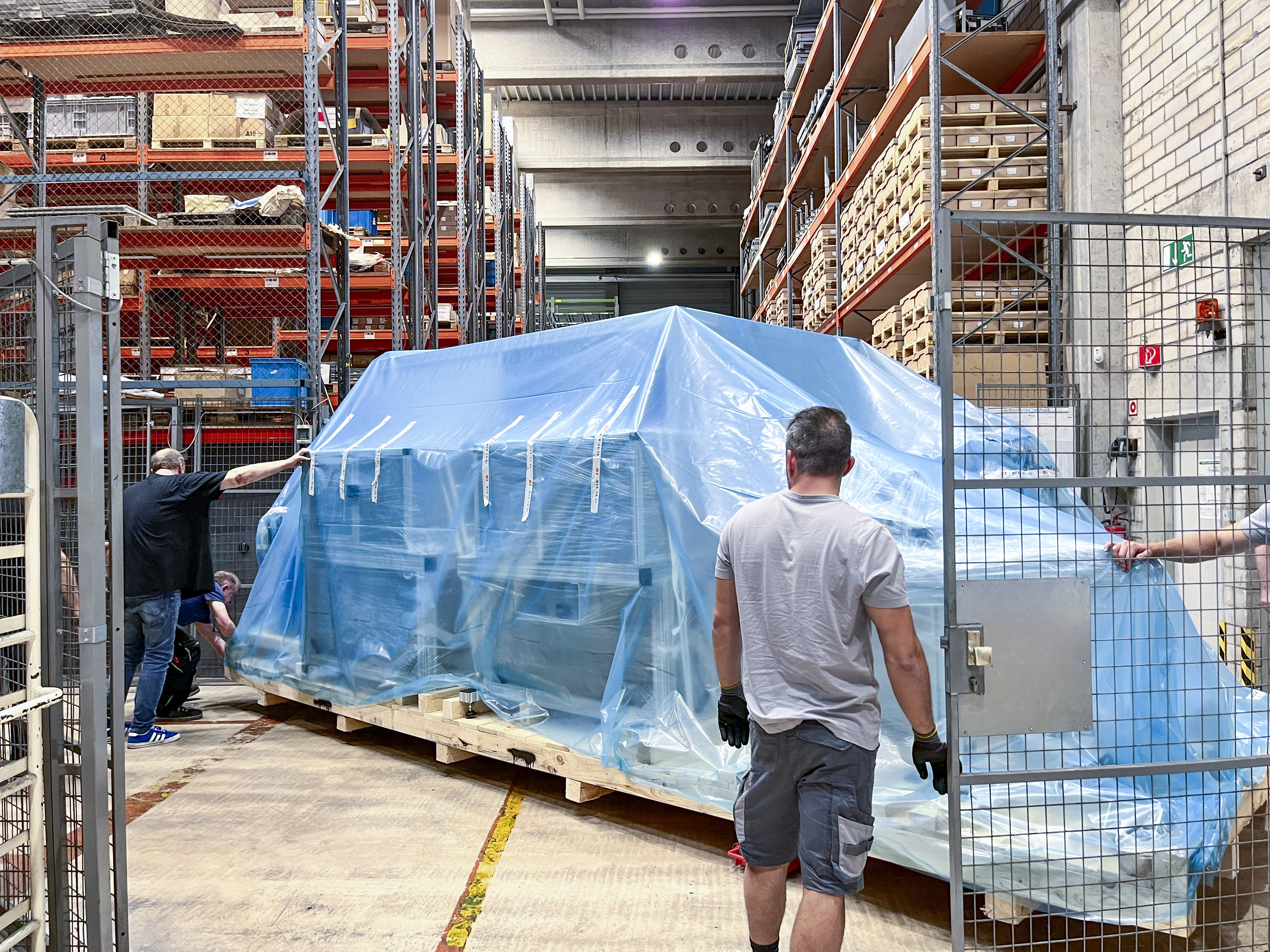Große Palette mit Teilen der Maschine wird im Lager rangiert; die Fracht ist mit blauer Plastikfolie geschützt