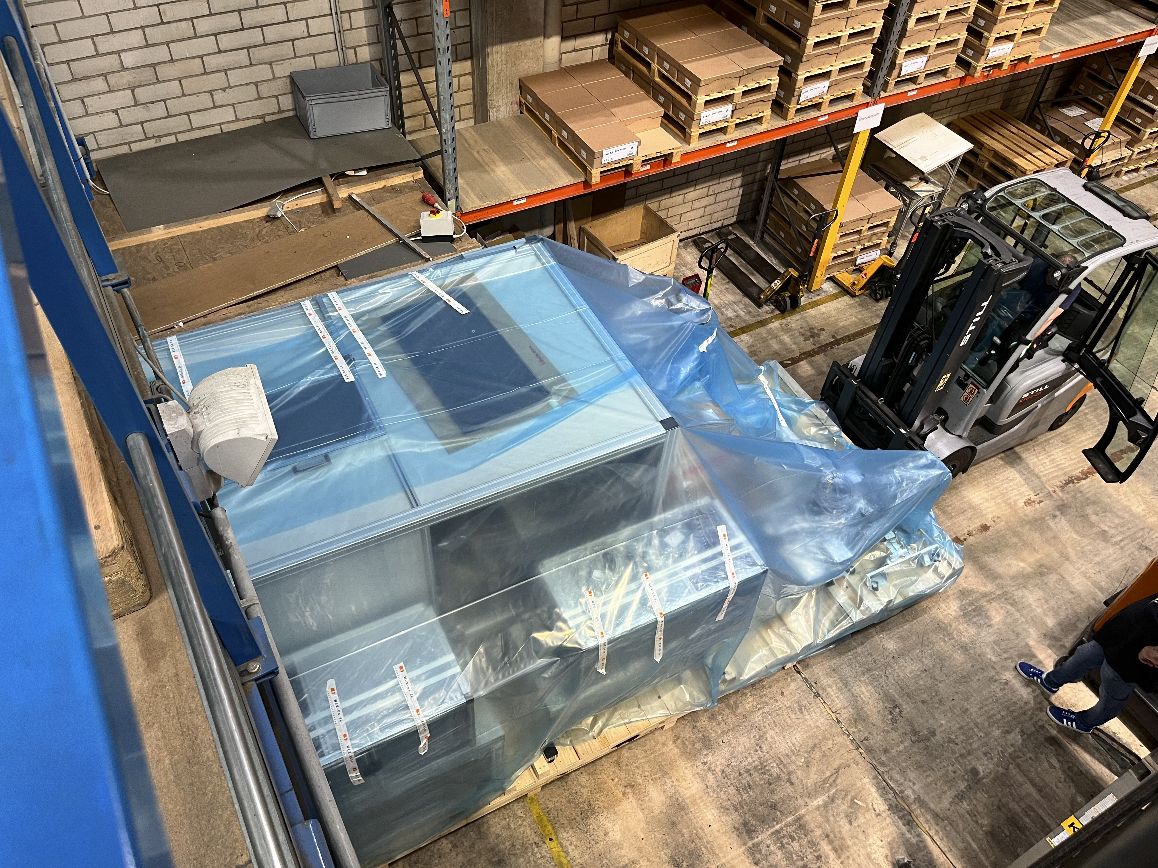Blick von oben: Eine große Palette mit Teilen der Maschine wird mit einem Gabelstapler ins Lager gefahren; die Fracht ist mit blauer Plastikfolie geschützt